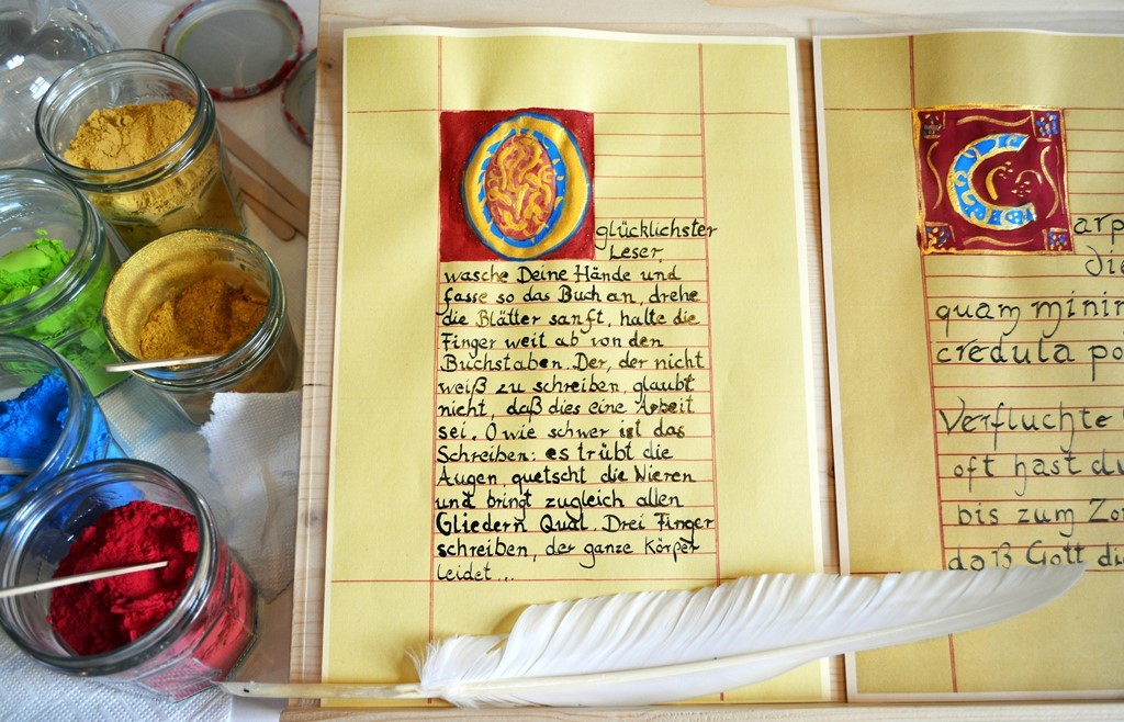Einer mittelalterlichen Buchseite nachempfundenes Blatt Papier mit kunstvoll gemalter Initale und handschriftlichem Text, eine Schreibfeder und links Farbpigmente in Gläsern.