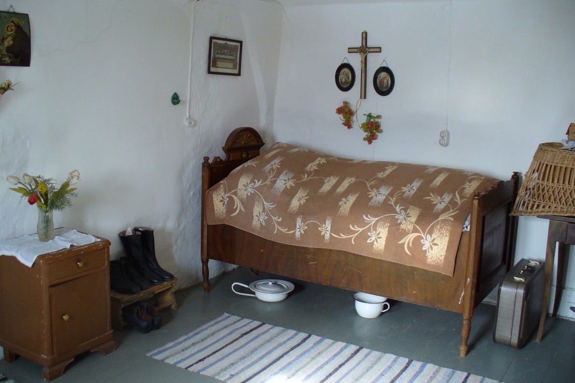 Schlafzimmer im Bauernhaus mit einfachster Einrichtung: ein schmales Bett, darüber ein Kruzifix und Heiligenbilder, unter dem Bett eine Bettpfanne. Links ein Nachtkästchen und drei paar Schuhe.
