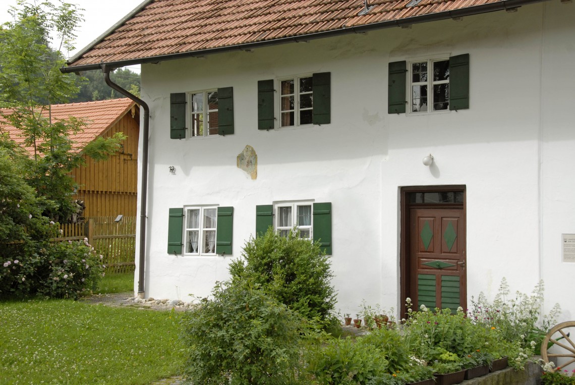 Das Weiß-Egger-Bauernhaus in Ellighofen. Weißes Haus mit grünen Fensterläden und braun-grüner Tür, davor Wiese und Büsche.