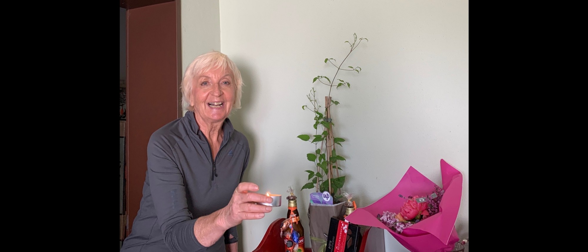 Ältere lachende Dame mit Kerze in der Hand, auf einem Tisch Geschenke und ein Blumenstrauß.