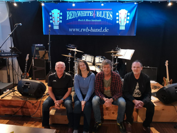 Die Bandmitglieder von &quot;Red, White and Blues&quot;, drei Männer und eine Frau, sitzen auf einer kleiner Holzbühne vor ihren Instrumenten und einem blauen Band-Banner.