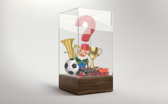 In einer Vitrine stehen neben einem großen Fragezeichen einige Objekte: eine Tuba, ein Fußball, ein Gartenzwerg, eine Modelleisenbahn und ein Pokal.