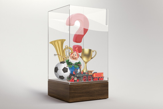 In einer Vitrine stehen neben einem großen Fragezeichen einige Objekte: eine Tuba, ein Fußball, ein Gartenzwerg, eine Modelleisenbahn und ein Pokal.