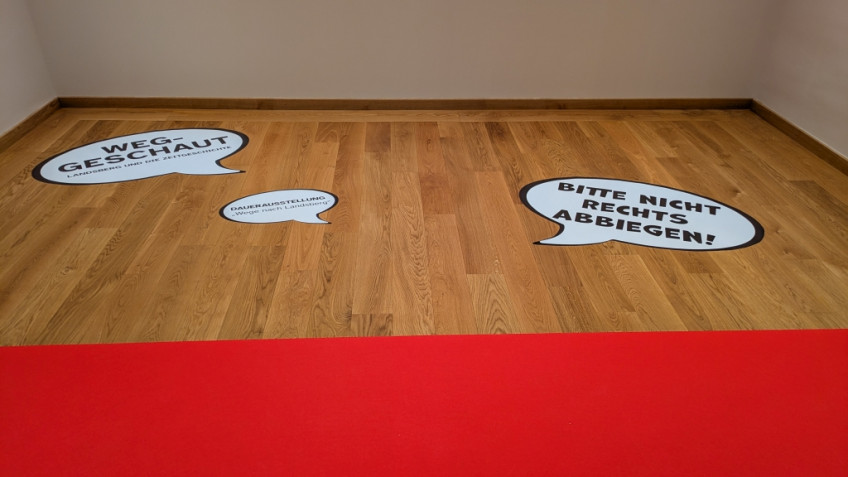 Drei Sprechblasen auf dem Parkettboden mit den Aufschriften "Weg-geschaut", "Dauerausstellung" und "Bitte nicht rechts abbiegen!"