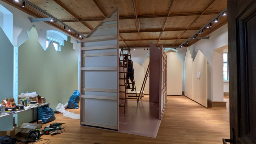 In einem Raum stehen farbige Stellwände in Form von Häusern entlang der Wände, in der Mitte des Raums wird ein freistehendes Haus aufgebaut.